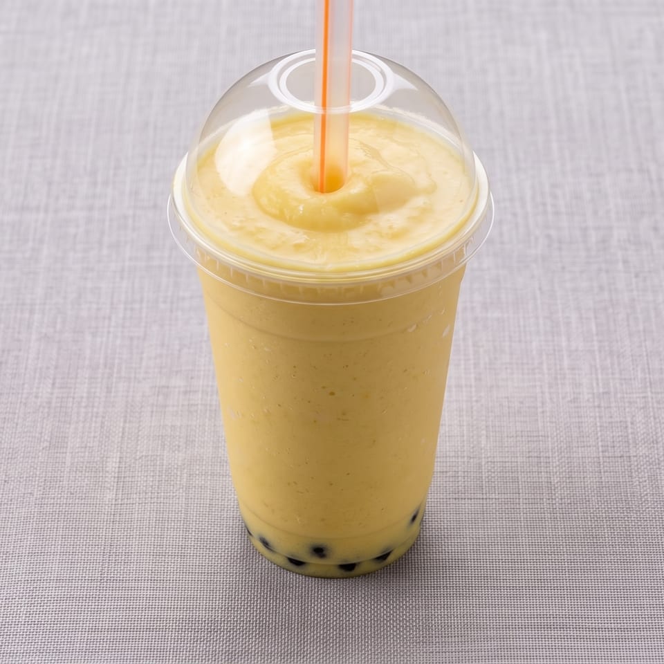 Yellow bubble tea smoothie with tapioca pearls in a clear lidded cup and orange straw on a fabric-covered surface.